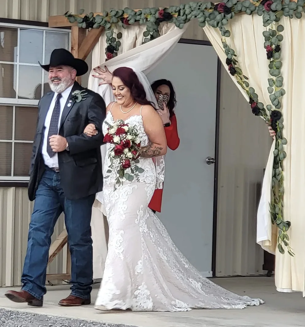 Bride in a lace gown with a red rose bouquet walking through the draped floral entrance with her father at Reverie at Diamond D in Normangee, Texas