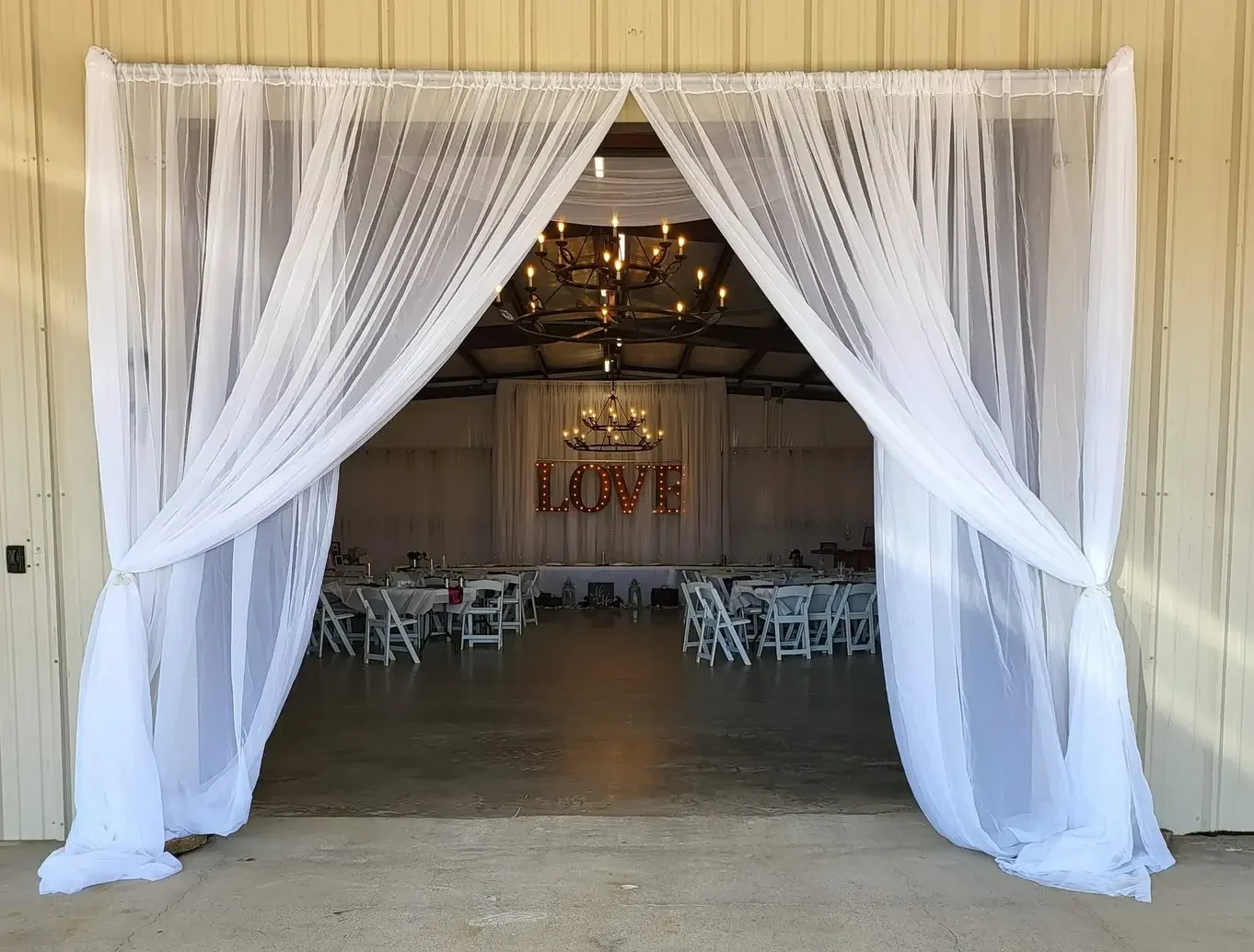Wide view through white drapery into the reception hall showing round guest tables, chandeliers and a lit LOVE marquee