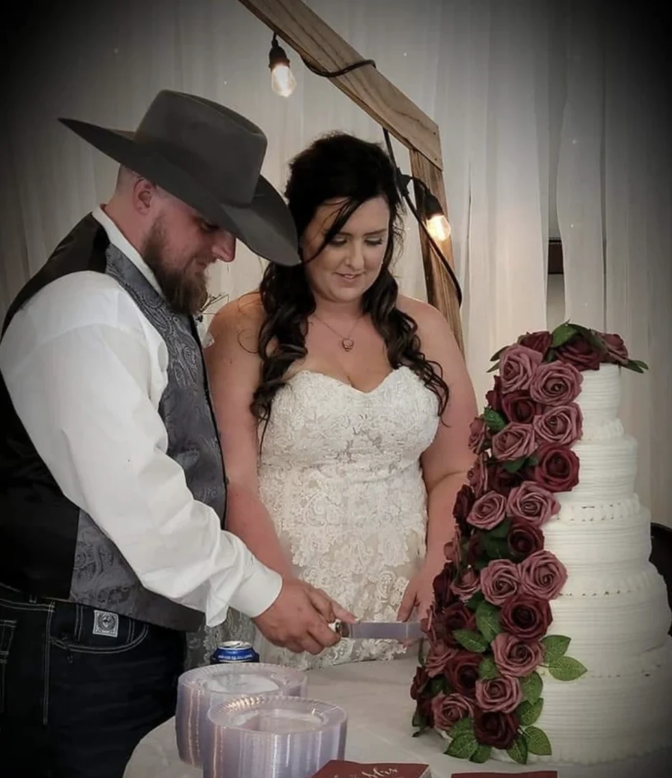 Cole and Emily cutting a tiered white wedding cake decorated with a cascade of burgundy roses in the reception hall
