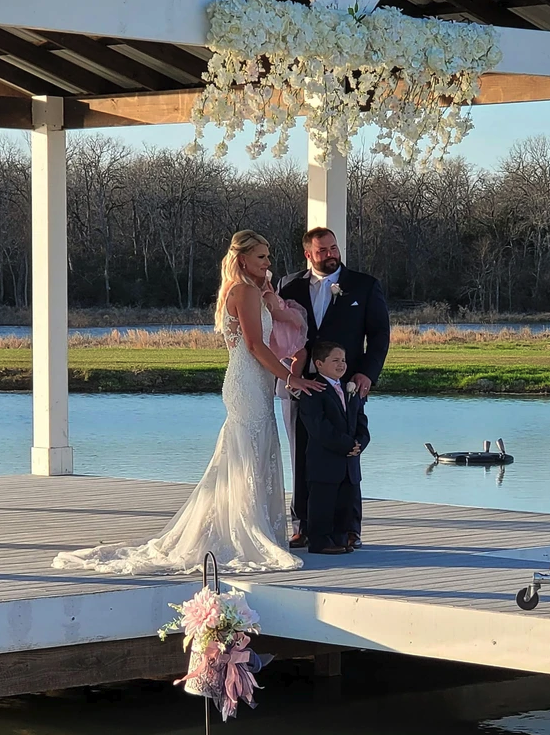 Bride, groom and son on the lakeside dock beneath the white floral chandelier