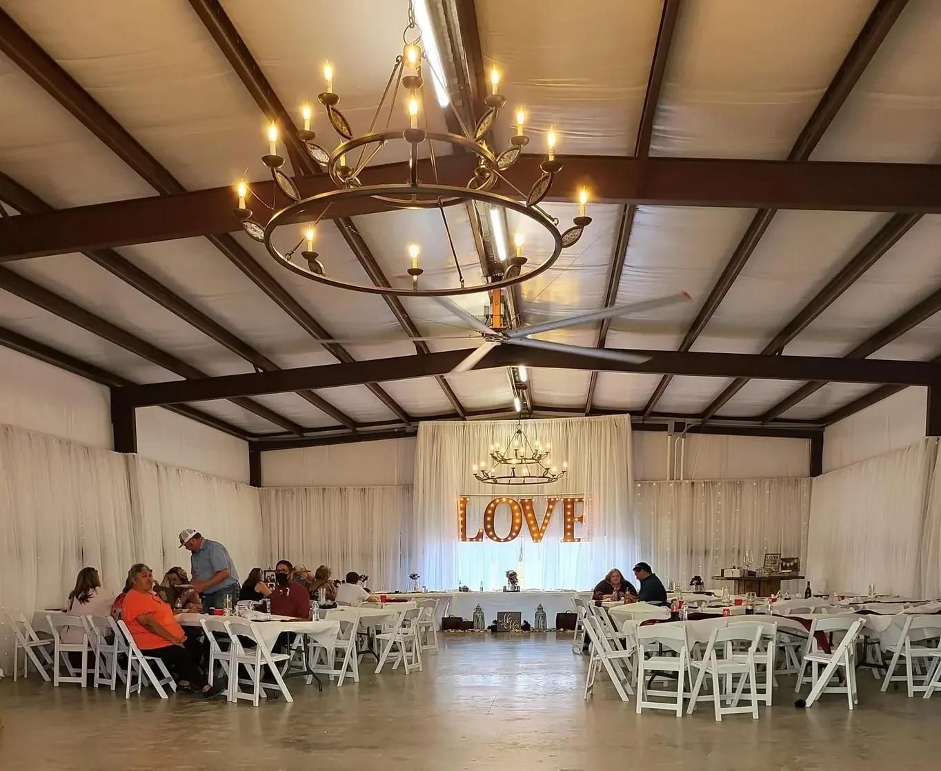 Guests seated at long white tables under the chandelier and LOVE marquee sign in the reception hall at Reverie at Diamond D