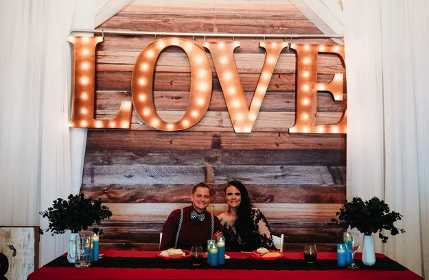 Newlyweds seated at the sweetheart table with red linens beneath the LOVE marquee backdrop
