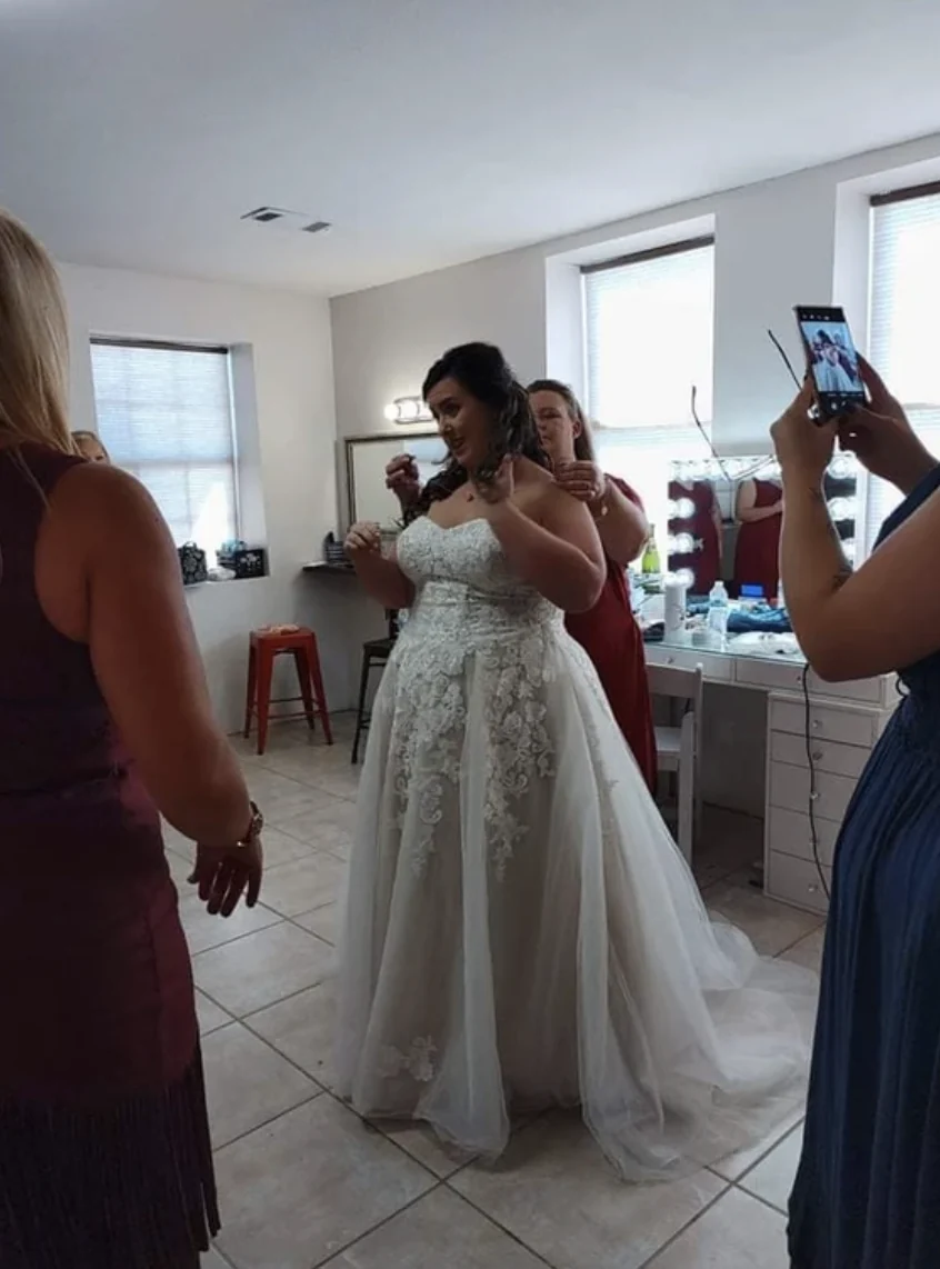 Bride putting on her necklace in the bridal suite with a lighted vanity mirror and bridesmaids nearby