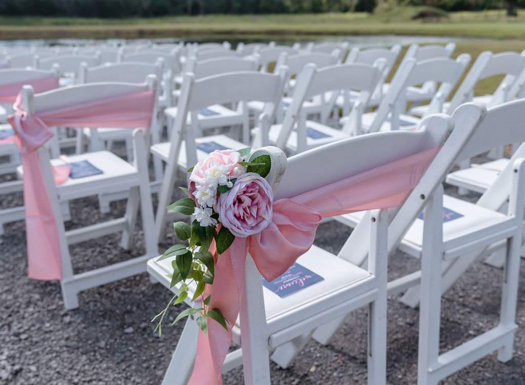 Close-up of a pink chiffon chair sash with roses lining the lakeside ceremony aisle