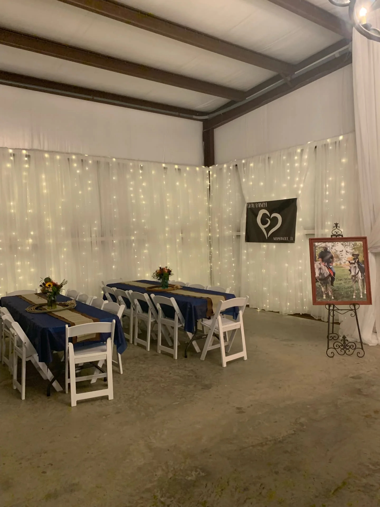 Two long guest tables with navy linens and floral centerpieces in front of walls draped with sheer fabric and warm string lights at the Love Like Bailin Therapy Farm event
