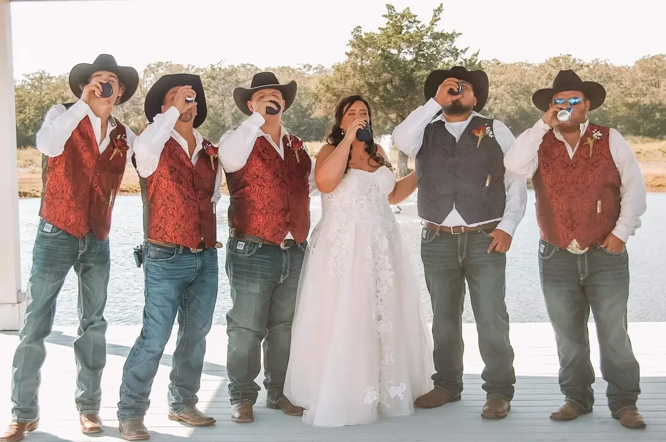 Bride in a lace gown drinking a toast alongside groomsmen in red vests on the lakeside dock