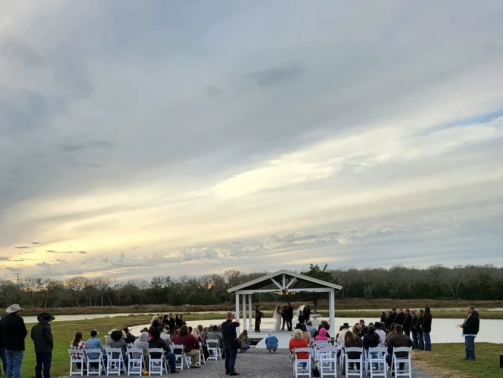 Wide sunset view of a wedding ceremony at the white lakeside pavilion with guests seated on the lawn at Reverie at Diamond D