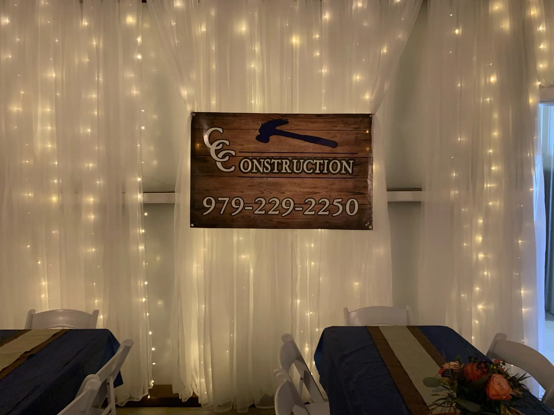 Wooden sponsor banner hung on a twinkle-light draped wall above navy-draped guest tables at the Love Like Bailin Therapy Farm event