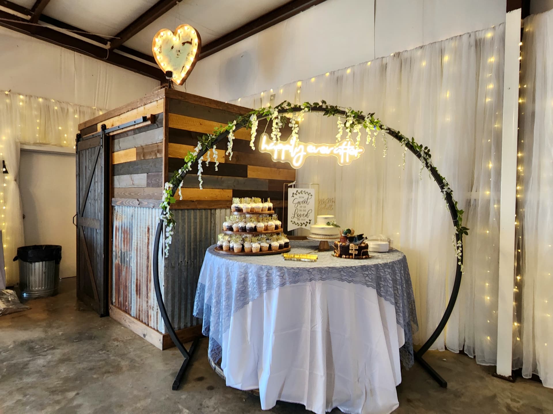 Cupcake tower and wedding cake under a circular greenery arch with happily ever after neon sign