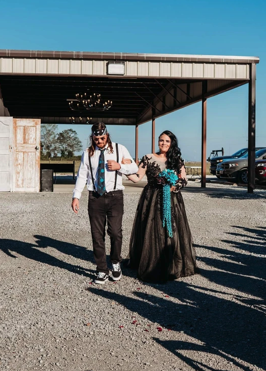 Bride in a black lace gown with turquoise bouquet being escorted from the covered pavilion