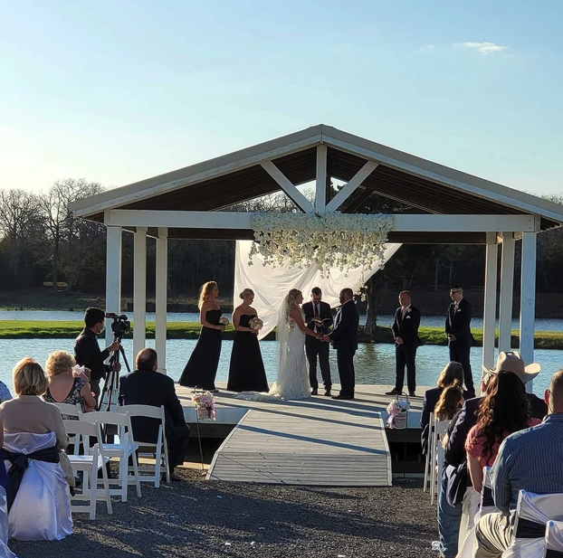 Couple exchanging vows beneath hanging florals as guests watch from the lakeside aisle