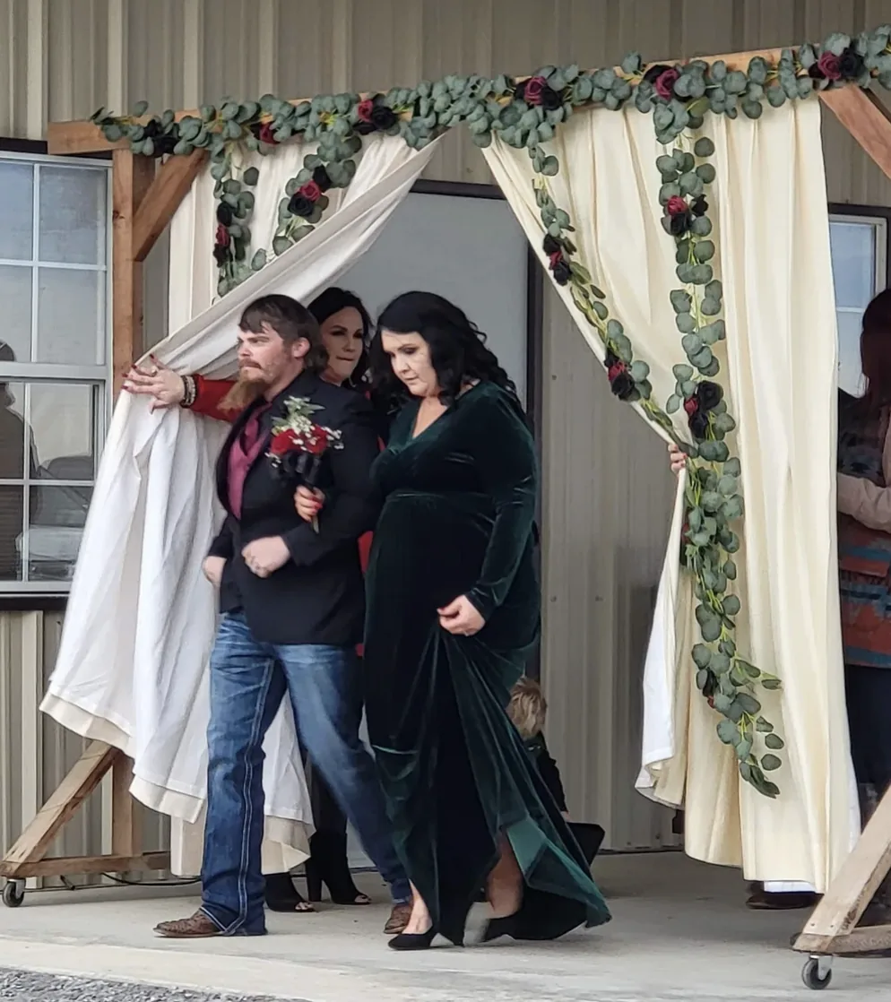 Family member in an emerald velvet gown escorted through the ivory draped floral entrance at a Normangee, Texas wedding