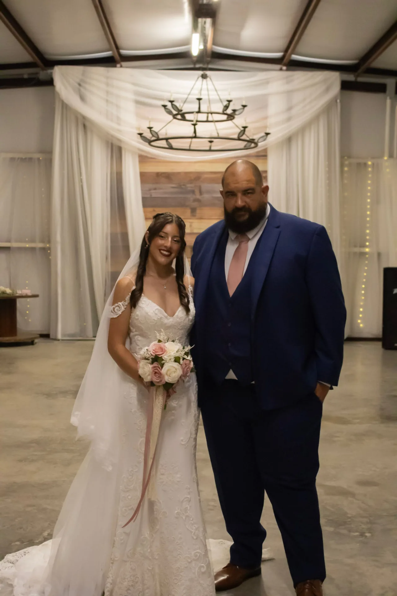 Bryanna in her lace gown with her father in front of the draped wood plank backdrop