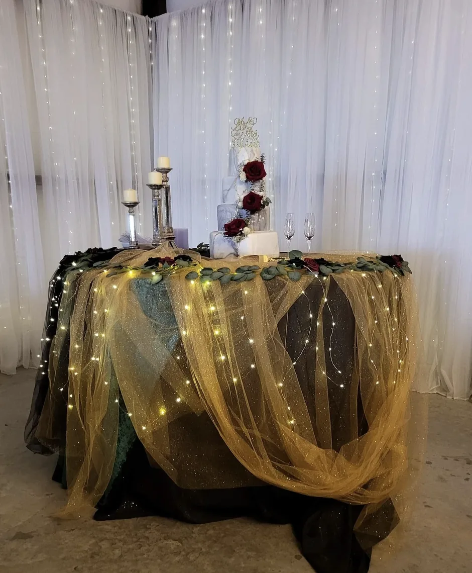 Three-tier wedding cake on a gold tulle draped table with twinkle lights, candles and burgundy roses at a Texas barn wedding venue