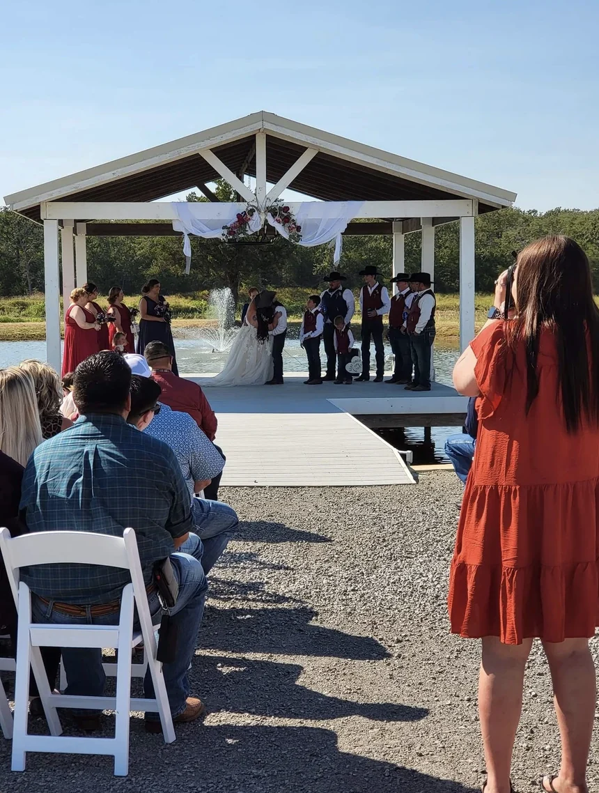 Guests seated on white chairs watching a wedding ceremony on the covered lakeside pavilion with the fountain running
