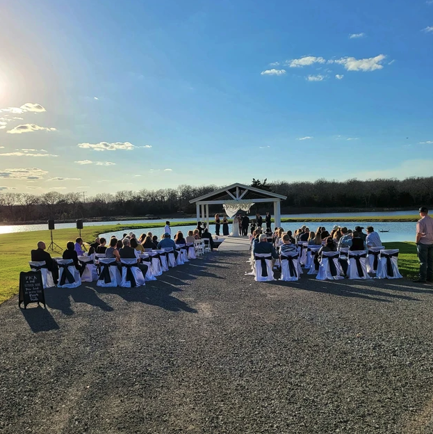 Golden hour lakeside ceremony with guests seated in black-sashed chairs facing the pavilion