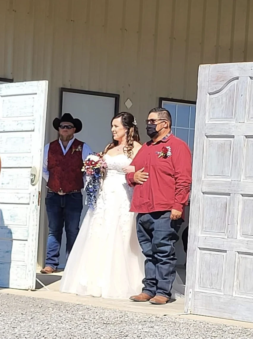 Emily and her escort framed by freestanding rustic white doors at the entrance to the outdoor wedding ceremony