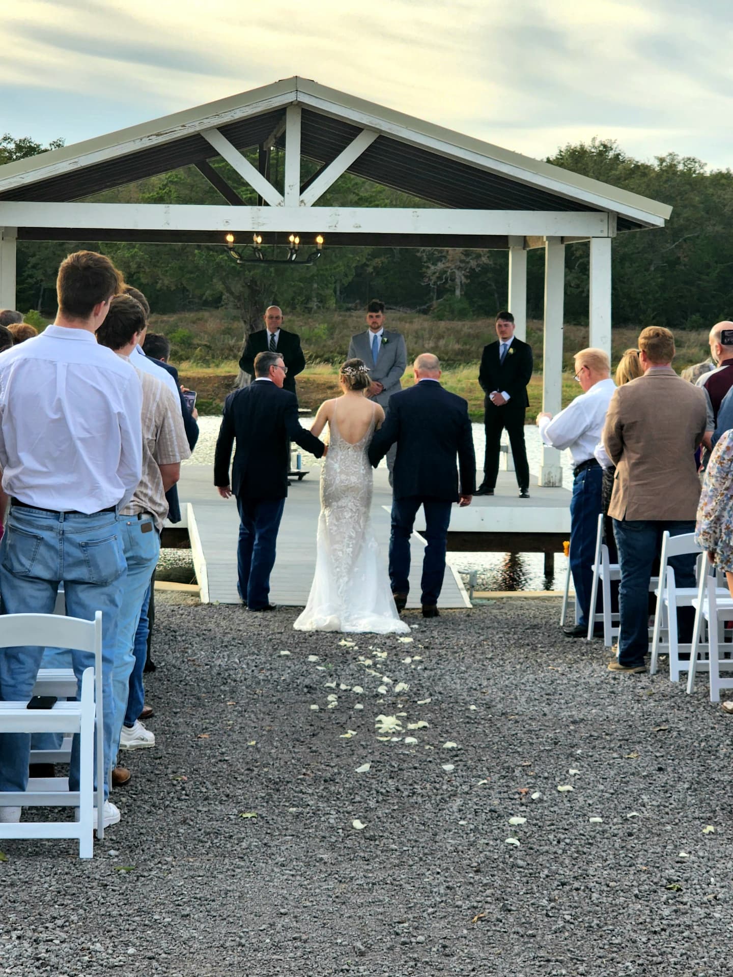 Guests standing as the bride walks the petal aisle toward the lakeside pavilion