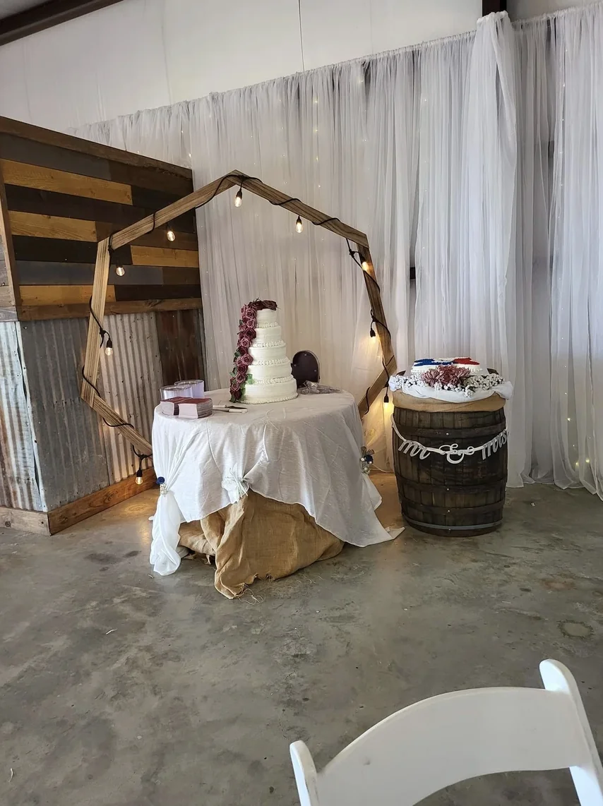 Tiered white wedding cake with burgundy roses beside a whiskey barrel and hexagon arch at a rustic Texas reception