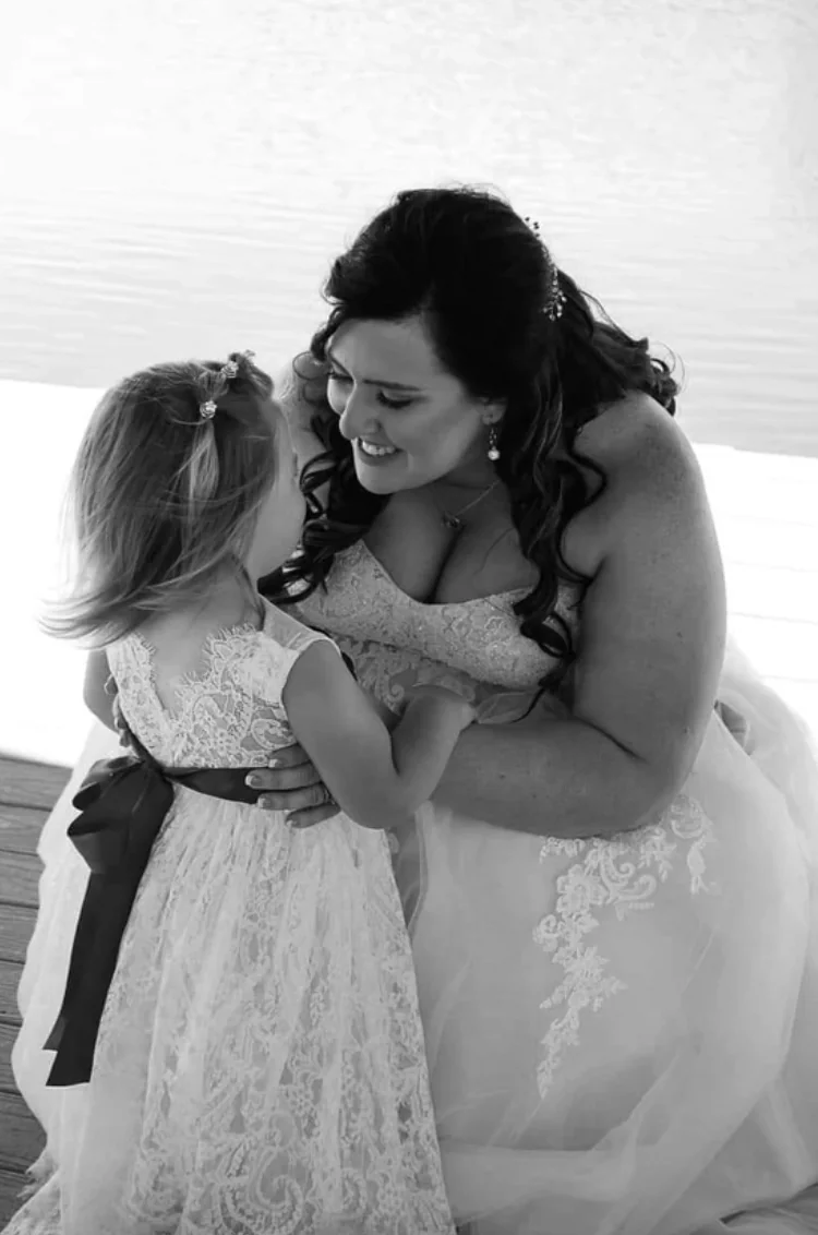 Black and white photo of the bride kneeling to hug the flower girl on the dock at a Texas country wedding venue
