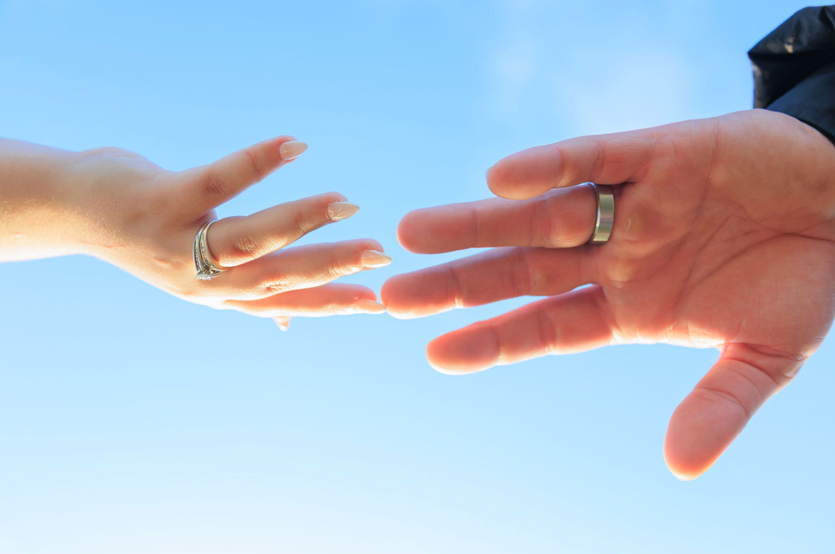 Jaelyn & Keelan's wedding at Reverie at Diamond D — the couple's hands reaching toward each other against a blue sky, rings visible