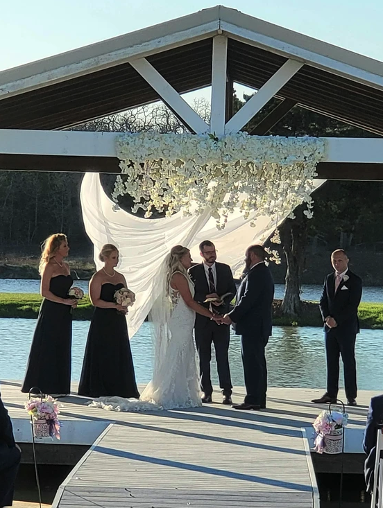 Couple exchanging vows under a hanging white floral arch on the lakeside pavilion