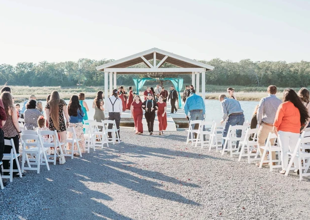Wide view of the lakeside ceremony aisle with guests seated in white chairs facing the pavilion