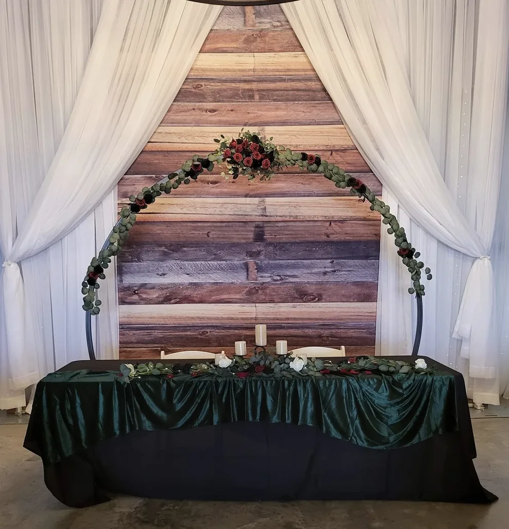 Sweetheart table with emerald velvet linens and a eucalyptus and rose arch in front of a reclaimed wood plank backdrop at Reverie at Diamond D