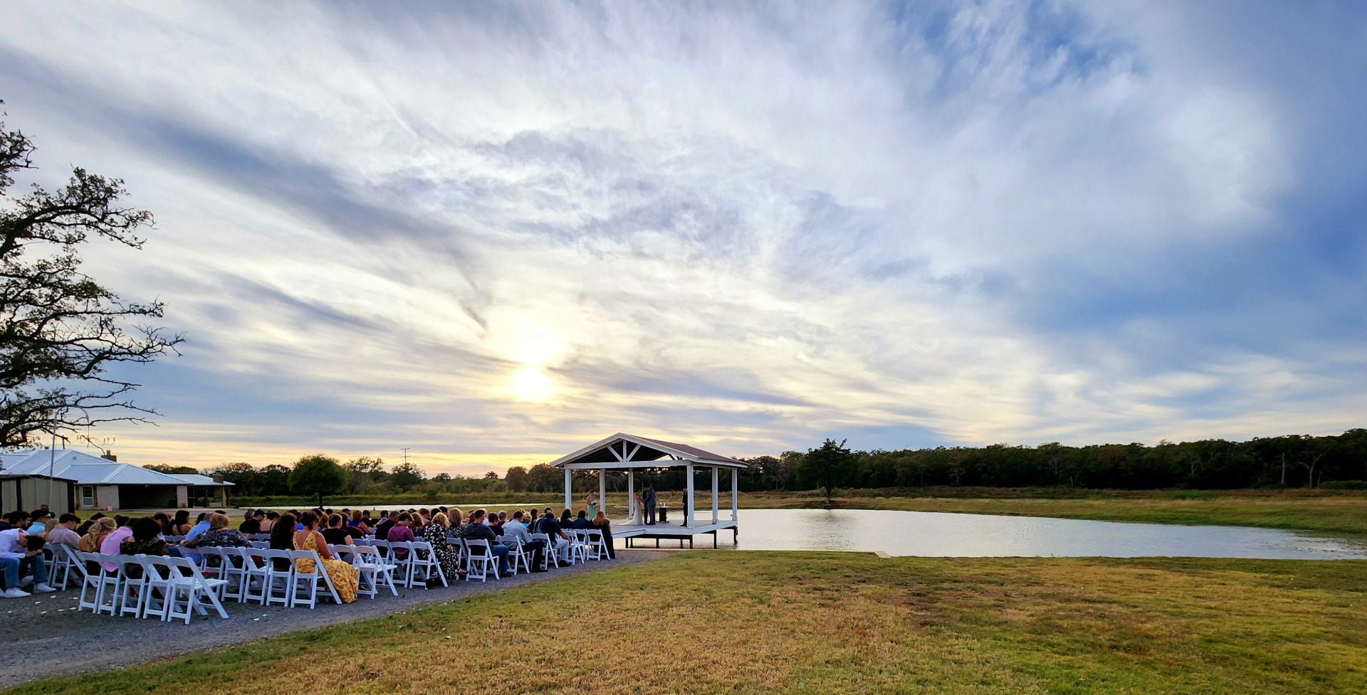 Sunset wedding ceremony at the lakeside pavilion with guests seated along the water at Reverie at Diamond D in Normangee, Texas