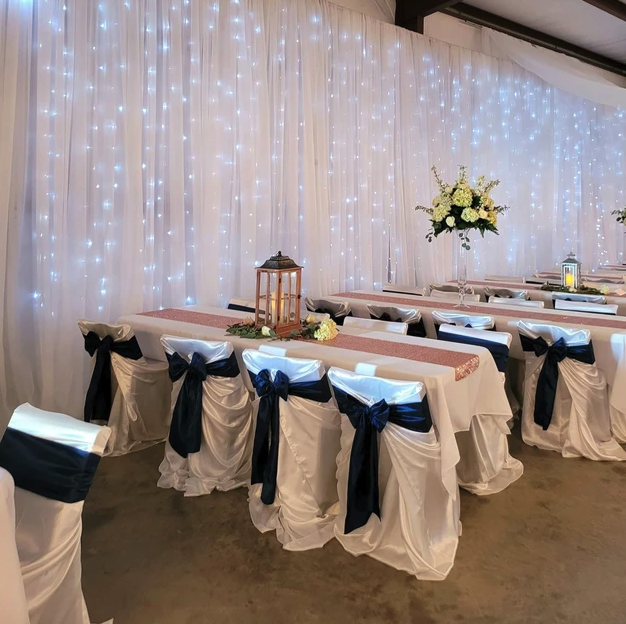 Head table with rose gold runners, lanterns and navy chair sashes in front of a twinkle-light backdrop