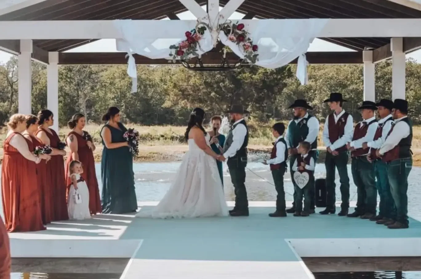Bride and groom holding hands during their vows beneath the lakeside pavilion with wedding party and flower girl