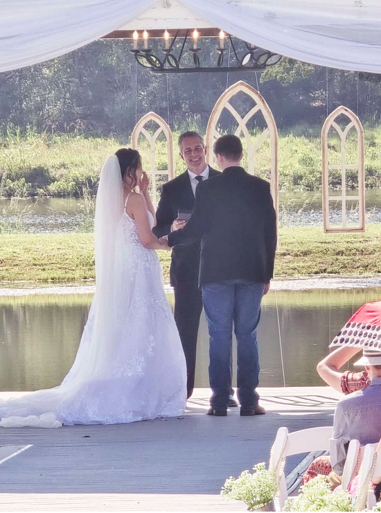 Bride and groom exchanging vows with the officiant beneath the pavilion chandelier and arched window backdrop