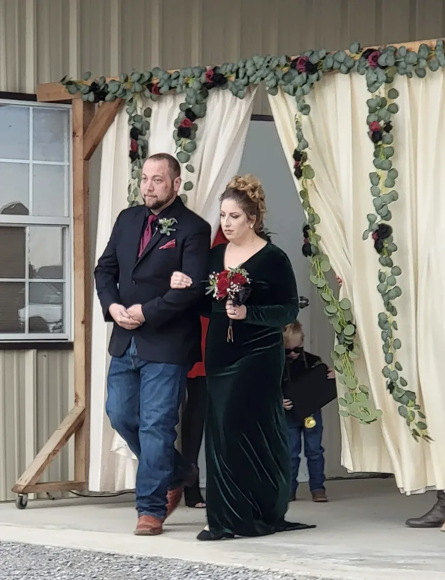 Groomsman and bridesmaid in an emerald velvet gown walking through the draped eucalyptus arch at Reverie at Diamond D