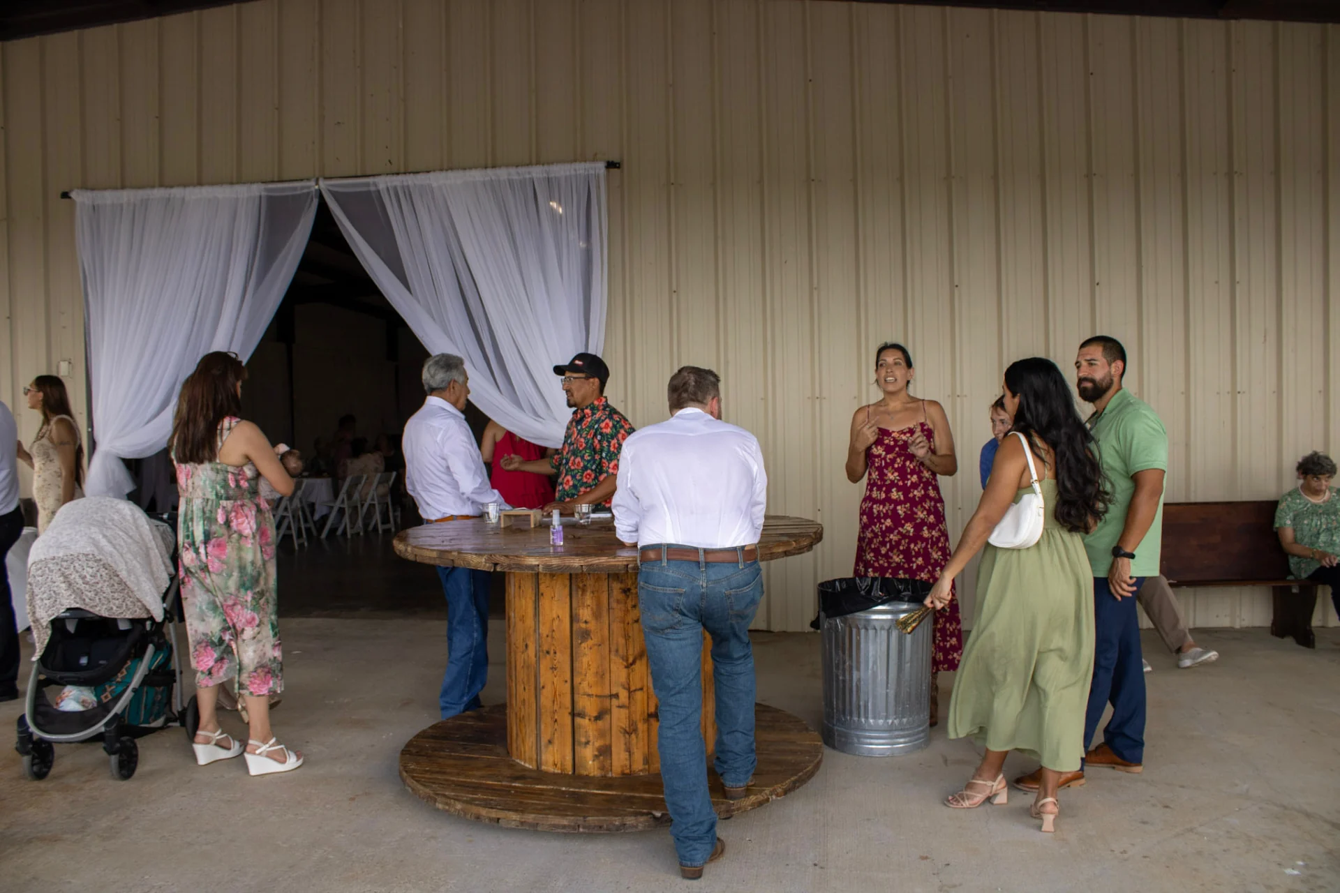 Wedding guests visiting around a wooden spool table on the covered patio outside the reception hall