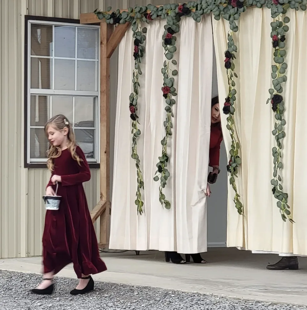 Flower girl in a burgundy velvet dress walking through the draped wedding ceremony entrance in Normangee, Texas