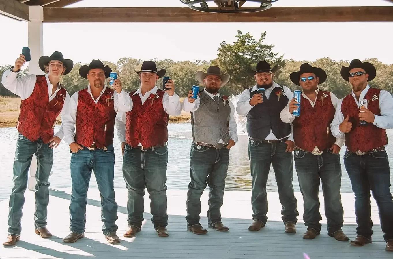 Groomsmen in cowboy hats holding drinks up in a toast on the covered dock beside the lake
