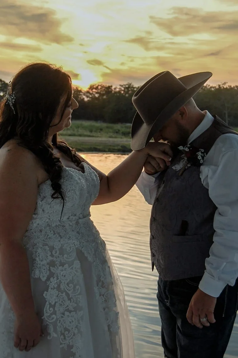 Groom in a cowboy hat kissing the bride's hand at sunset beside the lake at a Texas wedding venue