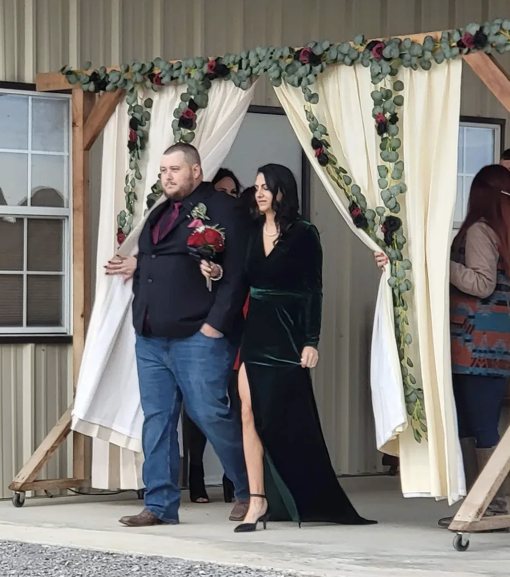 Bridal party walking through an ivory draped arch trimmed with eucalyptus and burgundy roses at a Normangee TX wedding venue