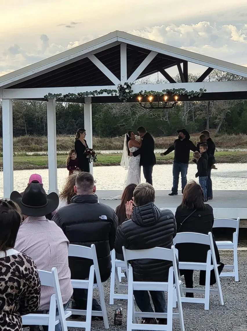 Bride and groom exchanging vows under the white lakeside pavilion with guests seated in white folding chairs at a Texas wedding venue