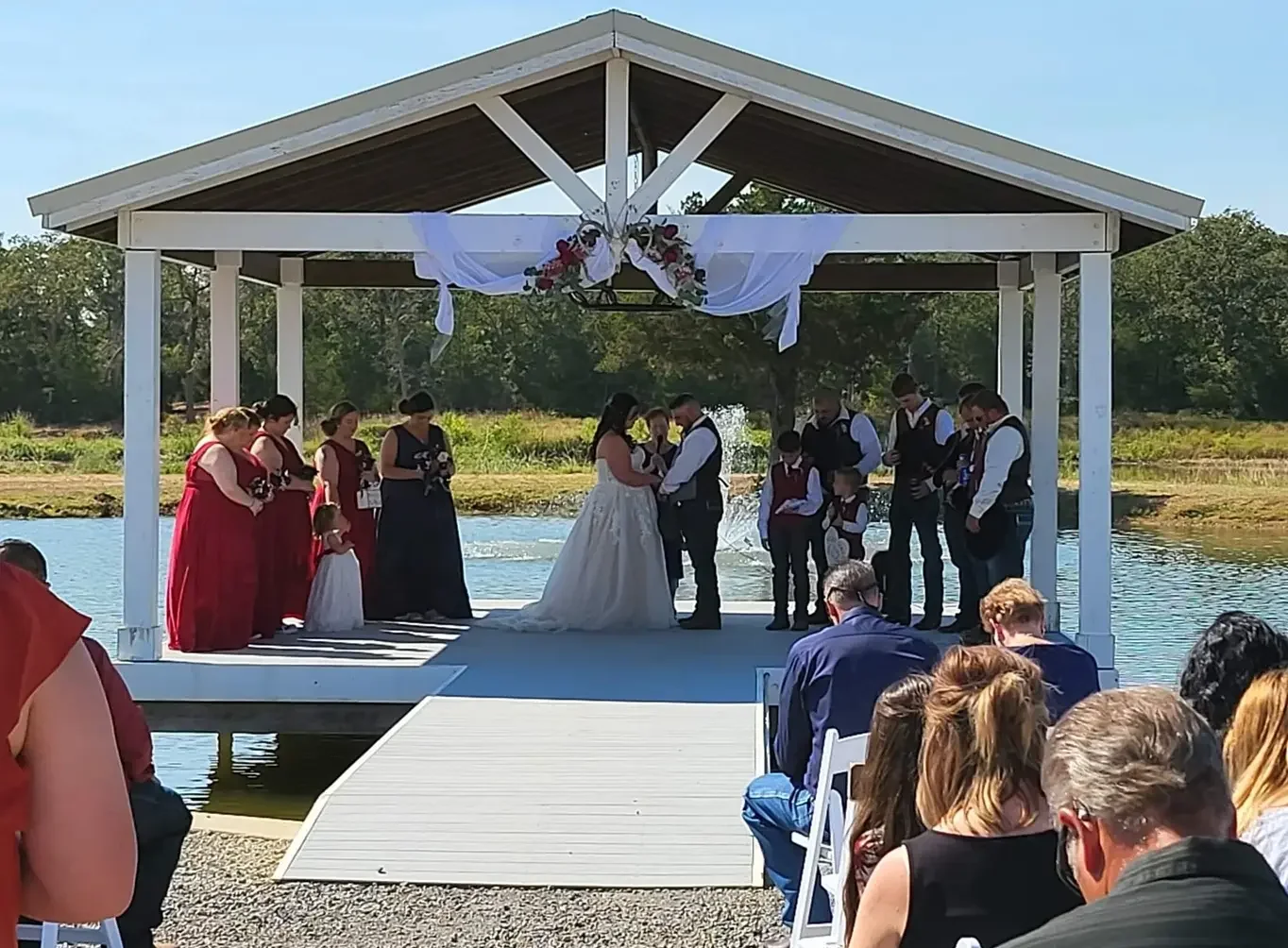 Wide view of guests seated for Cole and Emily's ceremony as the wedding party bows in prayer on the lakeside pavilion