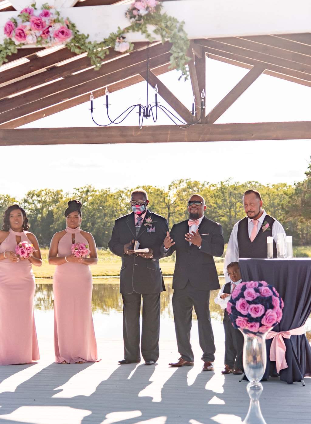 Wedding party and officiant standing beneath the timber pavilion beside the lake