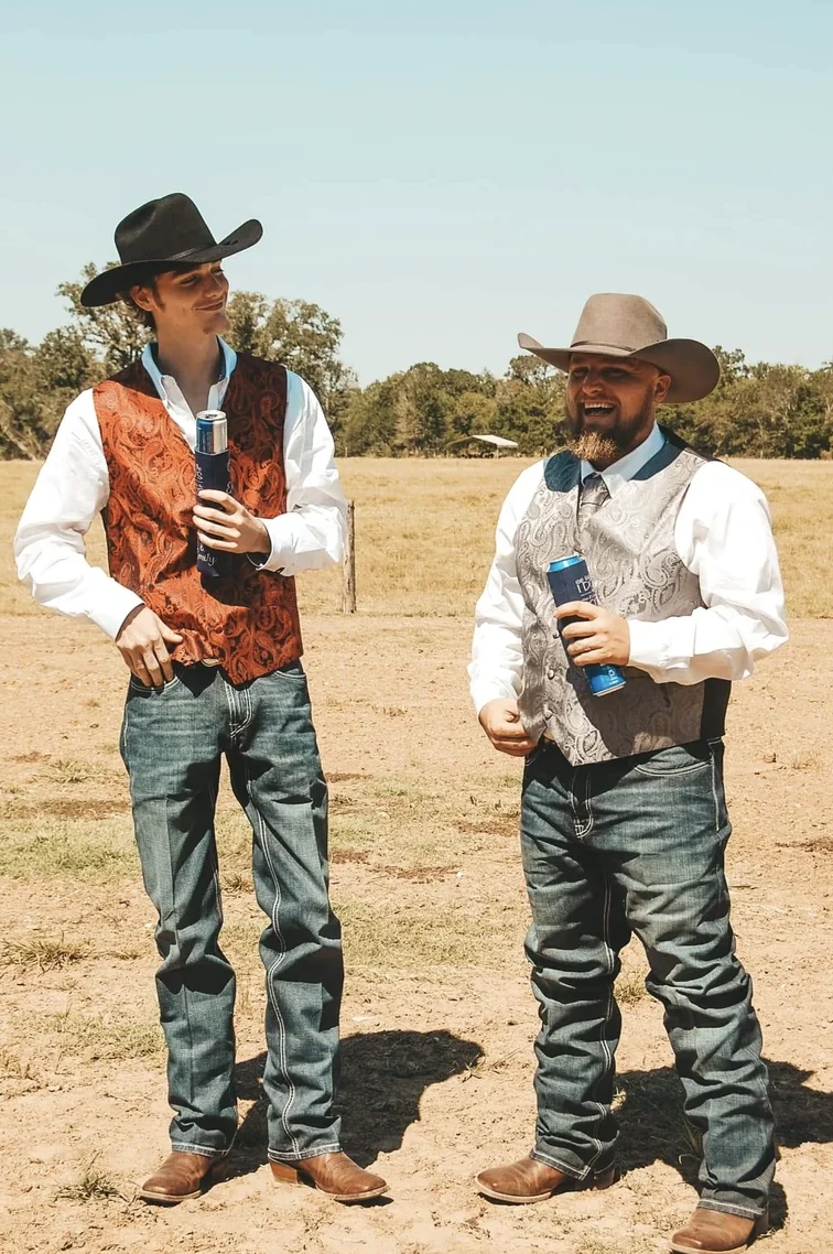 Two groomsmen in cowboy hats and paisley vests talking in an open ranch pasture before the wedding