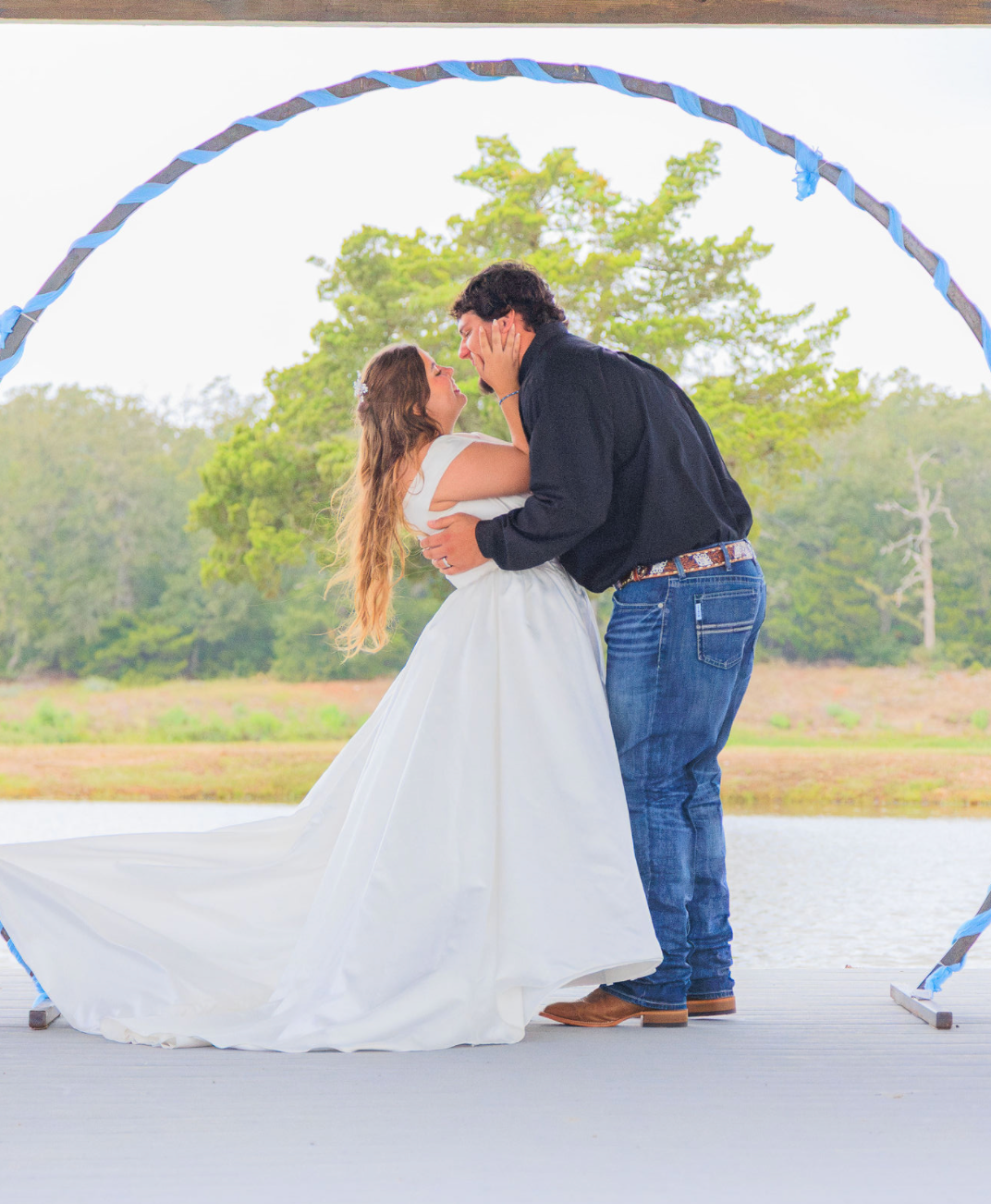 Jaelyn & Keelan's wedding at Reverie at Diamond D — the couple embracing on the lakeside pavilion dock beneath the blue-wrapped circular arch