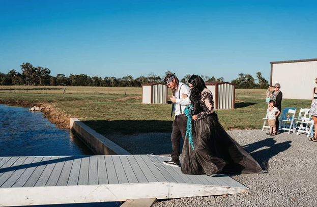 Bride in a black lace gown escorted toward the dock as guests look on