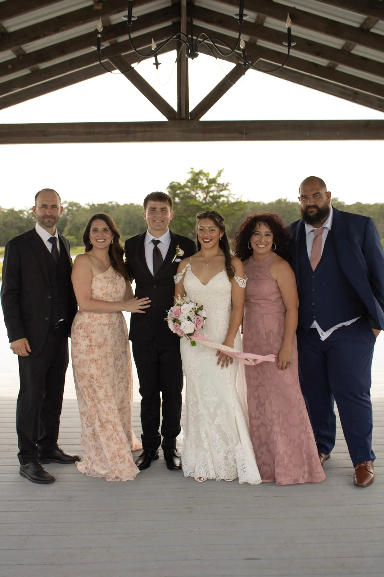 Newlyweds standing with their parents for a family portrait under the lakeside pavilion