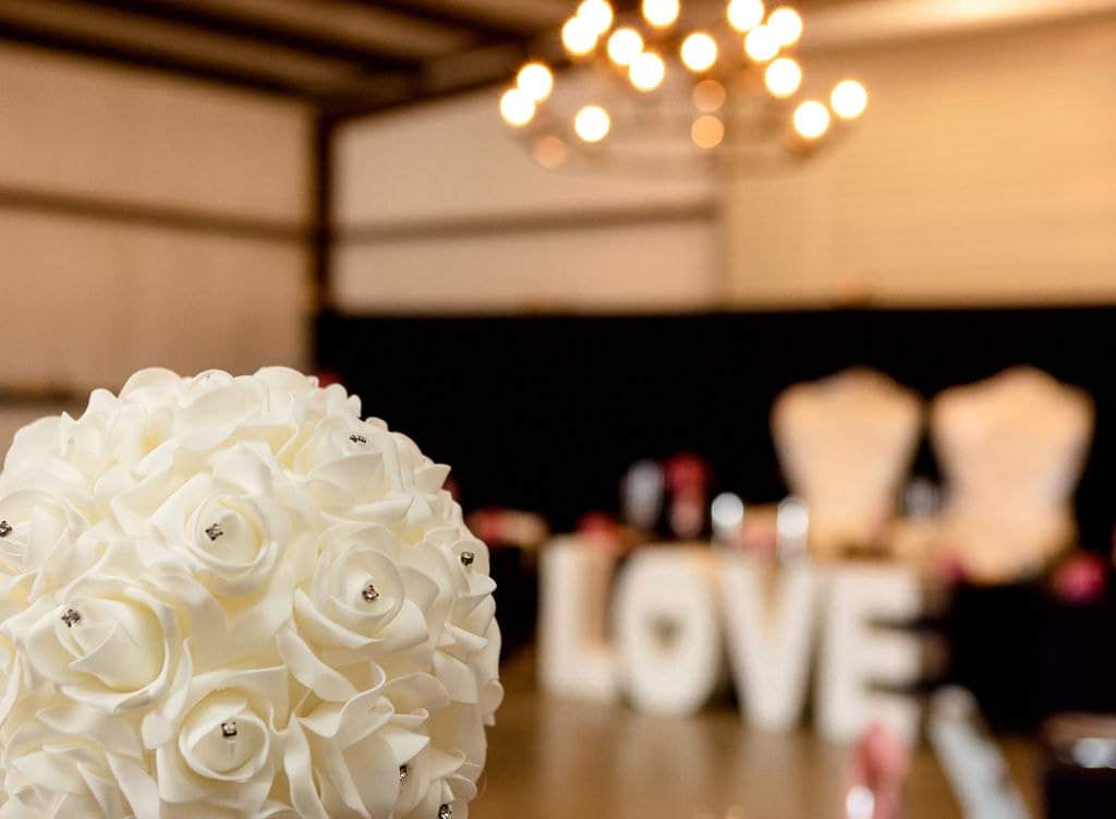 White rose bouquet in focus with the LOVE sign and head table glowing behind