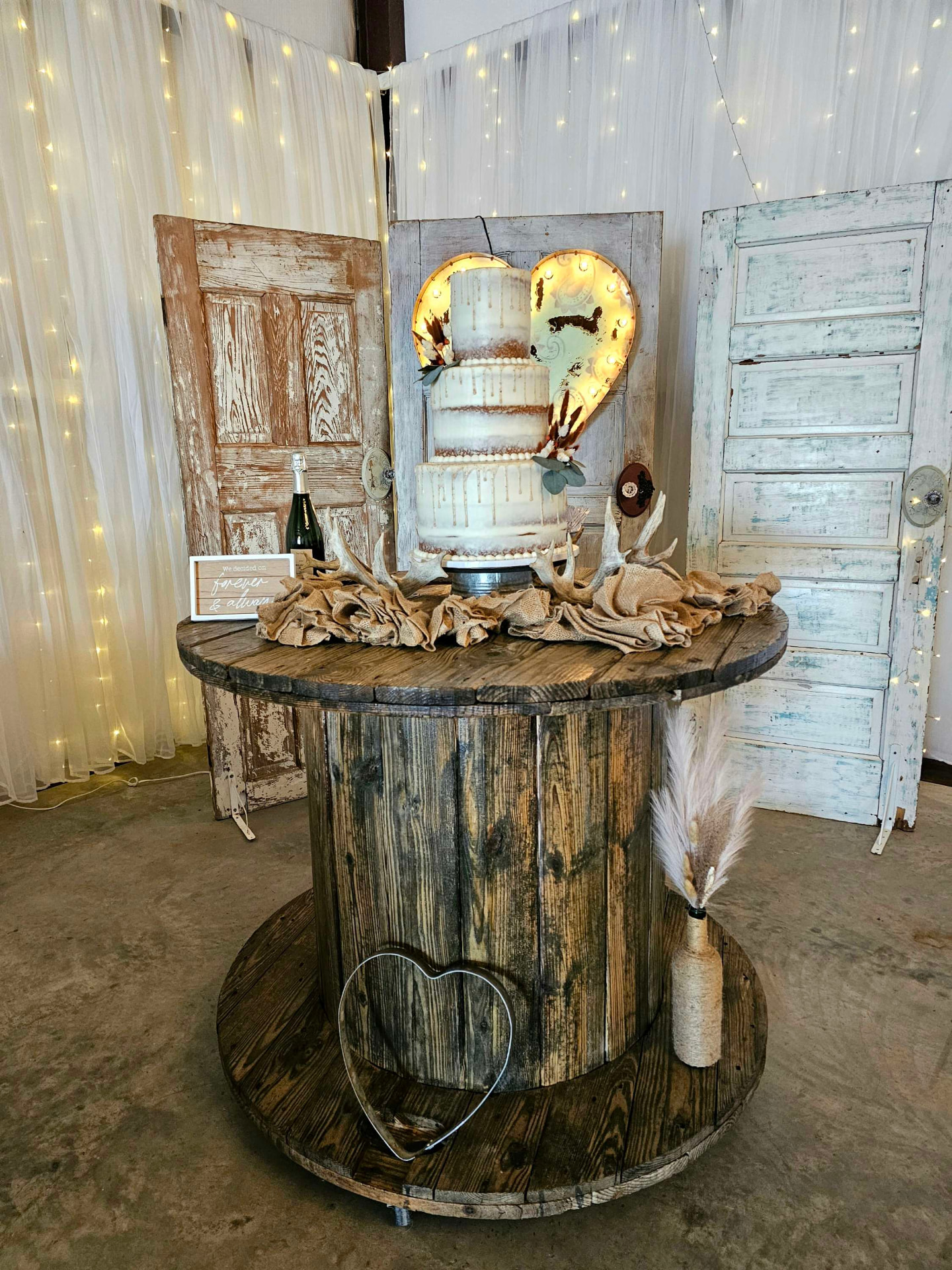 Three-tier naked wedding cake on a wooden spool table with antlers, vintage doors and a marquee heart at Reverie at Diamond D