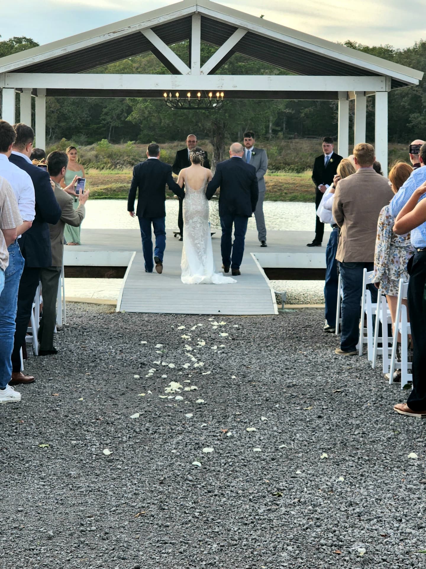 Bride walking the petal-lined aisle to the lakeside pavilion escorted by her fathers