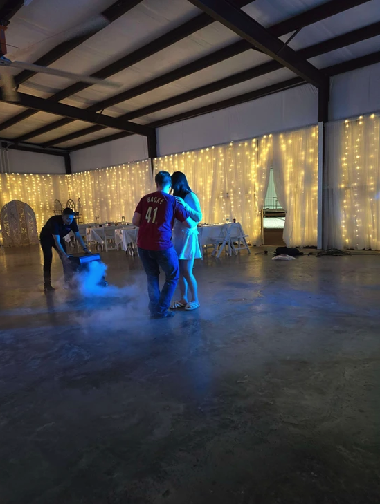 Wide view of a first dance with fog machine effects and twinkle-light backdrop in the barn