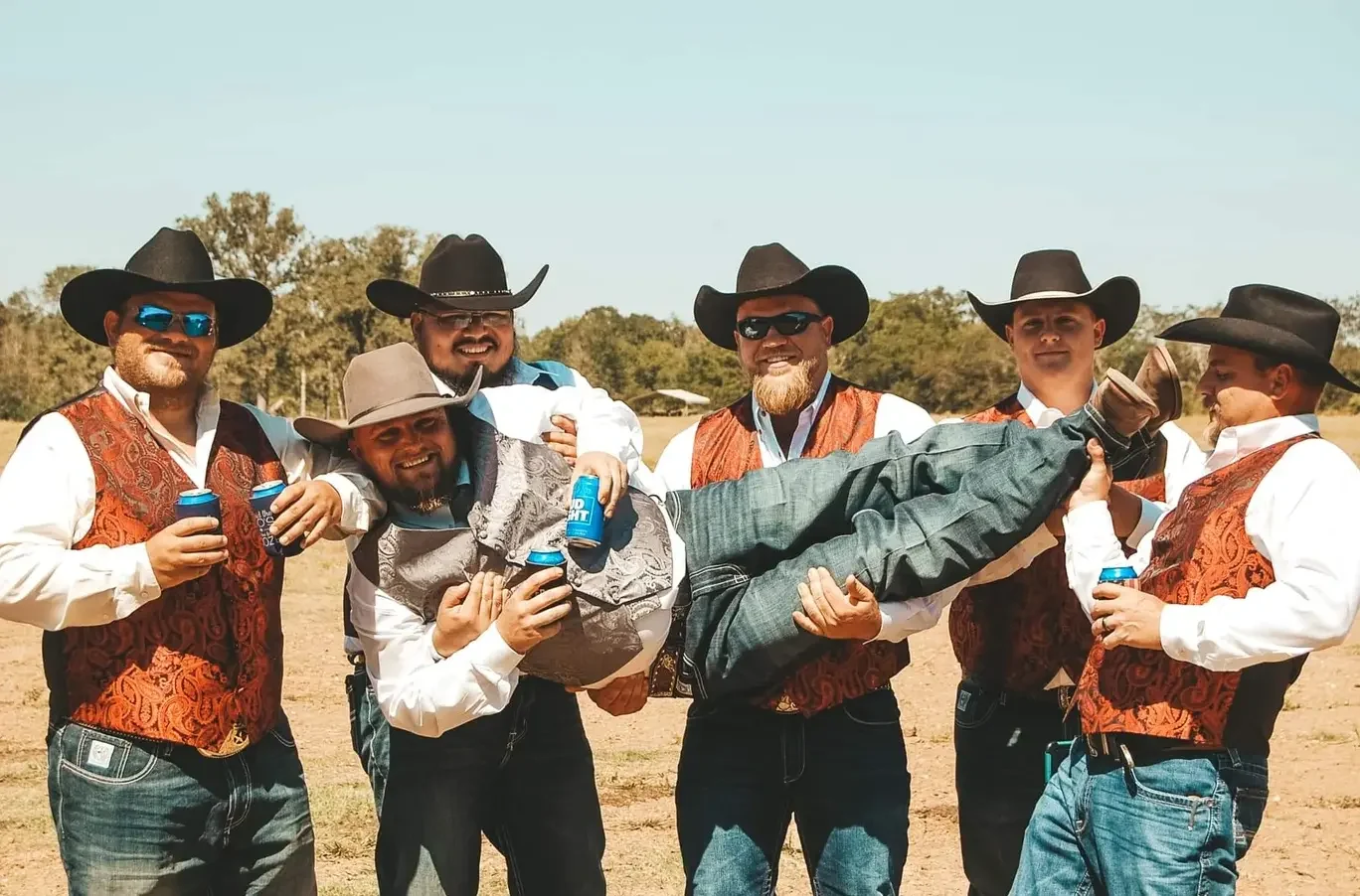 Groomsmen in cowboy hats and paisley vests lifting the groom in an open ranch field in Texas